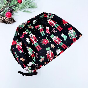 May include: Black scrub cap with a festive Christmas pattern. The design features red and green nutcrackers, poinsettias, gingerbread men, and stockings. The cap has a black elastic band for a secure fit. A perfect accessory for the holiday season.