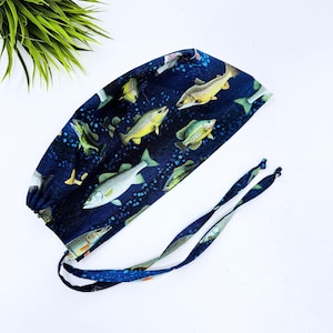 May include: A navy blue surgical scrub cap featuring a repeating pattern of various fish in shades of yellow, green, and white. The cap has long ties for securing and is set against a white background.