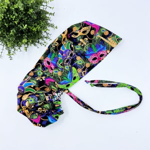 May include: A black surgical scrub cap with a vibrant Mardi Gras mask pattern. The cap features gold, green, purple, and pink masks, peacock feathers, and gold coin accents. The cap has a tie in the back for an adjustable fit.