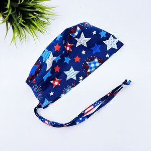 May include: A blue and red patterned surgical scrub hat with white stars and red and white checkered fireworks. The hat has two long ties.