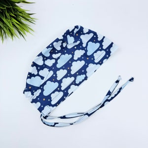 Puede incluir: Un gorro quirúrgico azul y blanco con estampado y lazos blancos. El estampado presenta nubes blancas sobre un fondo azul oscuro con pequeñas estrellas amarillas.