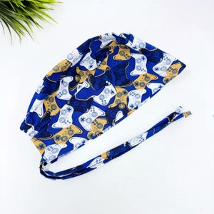 May include: A scrub cap featuring a vibrant blue background patterned with white and gold video game controller graphics. The head covering includes a gathered back section and two long fabric ties for securing the fit around the head.