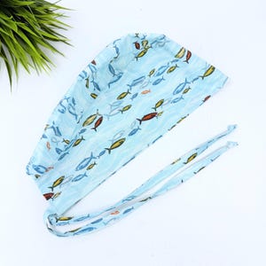 May include: A light blue scrub cap featuring a repeating pattern of small, colorful fish in shades of yellow, red, and dark blue. The cap has a rounded top, a wide band, and two long fabric ties extending from the back for a secure, adjustable fit.