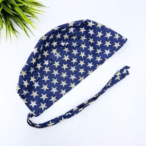 May include: A navy blue surgical scrub cap with a repeating pattern of white stars. The cap has a tie closure at the back and is made of a soft, flexible fabric. The cap is designed to be worn in a medical setting.