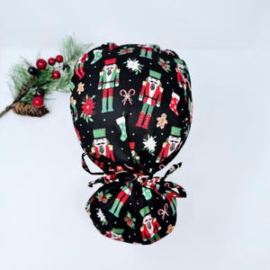 May include: A black scrub cap featuring a Christmas pattern with nutcrackers, gingerbread men, candy canes, and poinsettias in red, green, and white. The cap has a matching bow.