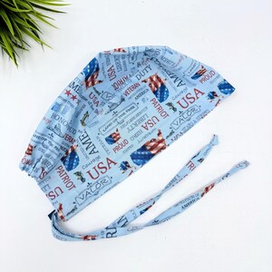 May include: A light blue surgical scrub cap with a patriotic USA theme. The cap features red, white, and blue imagery of the American flag and patriotic words like "USA", "Patriot", and "Valor". The cap has two long ties.