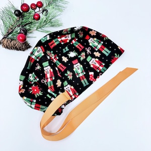 May include: A black scrub cap with a Christmas-themed pattern. The cap displays a repeating design of nutcrackers, gingerbread men, and festive accents in red, green, and white. A tan ribbon is attached for tying.