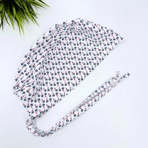 May include: A white scrub cap featuring a repeating pattern of small navy blue and red stars. The head covering has a rounded top, gathered fabric at the back, and two long fabric ties extending from the sides for securing the cap in place.