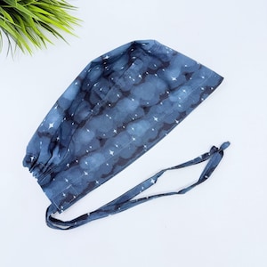 May include: A dark blue scrub cap featuring a repeating pattern of white four-pointed stars and soft, cloud-like shapes. The cap includes a gathered back section with integrated fabric ties for securing the fit, constructed from cotton fabric.