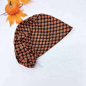 Puede incluir: Un gorro de quirófano negro con un patrón de caras de calabaza naranja. El gorro tiene un cordón blanco.