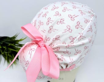 May include: White surgical scrub cap with a pink bow pattern and a large pink ribbon bow. The cap is designed to be worn in a medical setting. The cap is on a mannequin head.