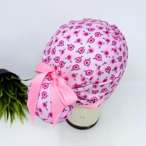 May include: A pink scrub cap with a floral pattern of small red and pink flowers. The cap has a matching pink ribbon bow on the back. The cap is designed to be worn in a medical setting.