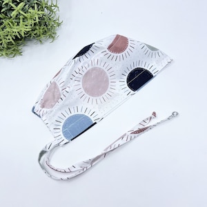 May include: A white surgical scrub cap with a colorful geometric pattern of pink, blue, and black circles with radiating lines. The cap has two long ties with the same pattern.