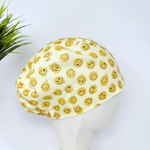 Puede incluir: Un gorro quirúrgico amarillo con un patrón repetido de caras sonrientes amarillas con detalles negros. El gorro está hecho de tela suave y está diseñado para cubrir la cabeza. El gorro está sobre una cabeza de maniquí blanca.