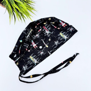 Op de afbeelding: Zwarte scrub-muts met een ruimtethema-print met kleurrijke raketten en planeten op een zwarte achtergrond. De muts heeft zwarte banden.