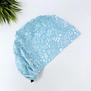 Op de afbeelding: Een lichtblauwe scrubcap met een wit bloemenpatroon. De cap heeft een geplooid rugpand en een donkerblauwe stropdas. De stof is bedrukt met bladeren, bloemen en andere botanische elementen.