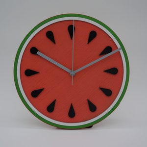 May include: A round watermelon-themed clock with a red face, black seed details, and a green and white border. The clock hands are gray. The clock is approximately 8 inches in diameter.