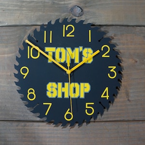 May include: Black circular clock with yellow numbers and hands. The clock is shaped like a saw blade and has the text "TOM'S SHOP" in yellow letters.