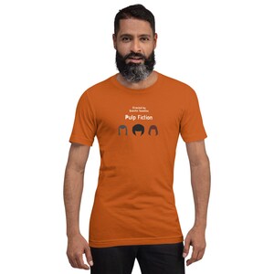 May include: Orange t-shirt with the text "Pulp Fiction" and the words "Directed by Quentin Tarantino" above it. The shirt features three stylized hair silhouettes. The person in the image is wearing the shirt.