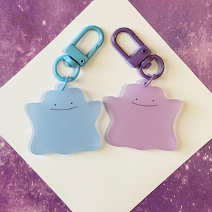 May include: Two keychains featuring a smiling, amorphous character in light blue and lavender. Each keychain has a matching colored clasp and ring. The background is a white diamond shape on a purple surface.