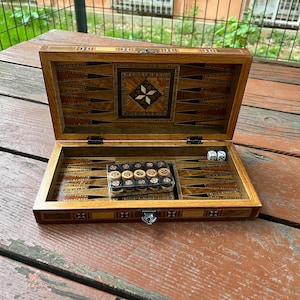 Könnte beinhalten: Offenes Backgammon-Set aus Holz mit eingelegten geometrischen Mustern. Das Spielbrett hat ein zentrales Sterndesign und enthält Würfel und Spielsteine. Das Set wird auf einer Holzoberfläche präsentiert.