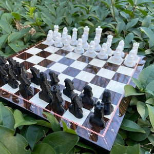 May include: A black and white chess set with the pieces arranged on a wooden chessboard. The board has a dark brown and light brown pattern. The pieces are made of plastic and have a matte finish.