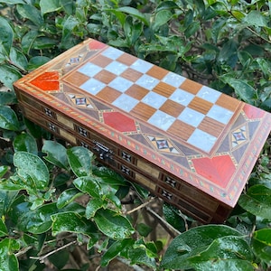 Könnte beinhalten: Ein rechteckiges Backgammon-Spielbrett aus Holz mit Schachbrettmuster. Das Brett hat Intarsien aus Holz und dekorative Akzente in Rot, Braun und Weiß. Das Brett ist geschlossen und hat einen Metallverschluss.