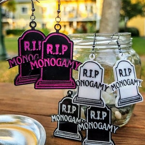 Peut inclure: Boucles d'oreilles en acrylique noir et rose en forme de pierres tombales avec le texte "R.I.P. Monogamie". Les boucles d'oreilles sont suspendues à une branche décorative argentée. D'autres boucles d'oreilles dans l'image sont blanches et noires.