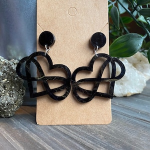 May include: Black heart-shaped earrings with an intricate, interwoven design. The earrings are suspended from small, round black studs. The design features a gold-colored marbling effect within the black acrylic. The earrings are displayed on a brown card.