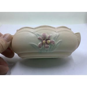 May include: A cream-colored ceramic planter with a scalloped rim and handles. The planter features a raised floral design with pink and yellow flowers and green leaves. The planter is a decorative item.
