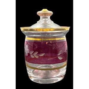 May include: A clear glass jar with a lid, featuring a decorative design. The jar has a band of deep red with etched floral patterns and gold trim. The lid is clear with a gold top. The jar is likely used for storing small items.