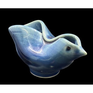 May include: A blue ceramic bowl with a unique, flowing design. The bowl has a rounded base and a rim that curves upwards, creating an organic shape. The glossy finish reflects light, highlighting the bowl's form. The bowl is a decorative item.