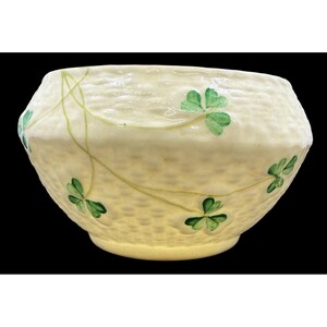 May include: Cream-colored ceramic bowl with a textured surface and green shamrock designs. The bowl has a wide, open top and a slightly flared base. Green vines with shamrocks are painted around the bowl.