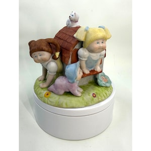 May include: A ceramic music box featuring a house with a brown roof and three doll-like figures. One figure is crawling, another is peering out of the house, and the third is leaning over the house. A small white dog sits on the roof.