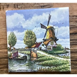 Può includere: Piastrella quadrata in ceramica raffigurante un pittoresco paesaggio olandese. L'immagine mostra un tradizionale mulino a vento con pale rotanti, un ponte, un fiume e case con tetti rossi. Il cielo è blu con nuvole bianche.
