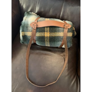 May include: A rolled-up green and yellow plaid blanket with brown leather straps and a handle. The blanket is secured with leather straps and buckles, and has a leather handle for carrying. The blanket is on a brown leather surface.