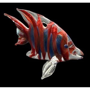 Könnte beinhalten: Eine handgefertigte Glasfischskulptur mit lebendigem Design. Der Fisch hat rote und blaue Streifen auf einem transparenten Glaskörper. Er hat eine spitze Schnauze, Flossen und ein kleines blaues Auge. Die Skulptur ist vor schwarzem Hintergrund.
