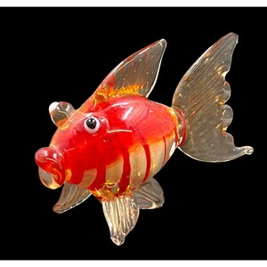 Könnte beinhalten: Eine handgefertigte Glas-Goldfisch-Figur. Der Fisch hat einen leuchtend roten Körper mit klaren, gestreiften Akzenten und Flossen. Die Augen sind schwarz und weiß. Die Figur ist vor einem schwarzen Hintergrund platziert.