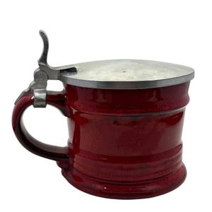 May include: A burgundy ceramic beer stein with a metal hinged lid and handle. The stein has a glossy finish and a ridged design around the base. The handle and lid hardware are silver-toned.