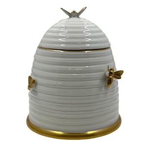 May include: White ceramic beehive-shaped jar with a lid, accented with gold trim and bee embellishments. The jar has a textured, ribbed design and a gold bee on top of the lid. A decorative item for the kitchen or home.