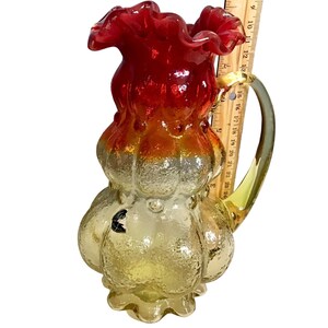 May include: A decorative glass pitcher with a ruffled red rim, transitioning to orange and yellow. The pitcher has a textured surface and a clear handle. A ruler is placed next to the pitcher, showing measurements in inches.