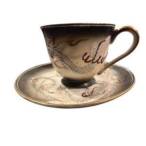 May include: A ceramic teacup and saucer set. The cup and saucer are gray with a gold trim. A white dragon design is featured on both pieces. The cup has a handle and the word "Wu" in red. The set is on a white background.