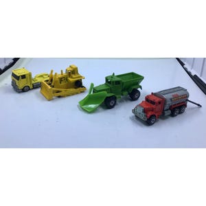 May include: A collection of toy construction vehicles, including a yellow dump truck, a yellow bulldozer, a green dump truck, and a red tanker truck. The toys are arranged on a white surface.