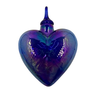 Puede incluir: Una botella de vidrio en forma de corazón con un tapón largo y delgado. La botella tiene un acabado azul oscuro iridiscente con toques de morado y verde. El diseño es liso y reflectante, adecuado para uso decorativo.