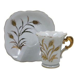 May include: White teacup and saucer set with a gold wheat design. The cup has a gold handle and rim, and the saucer has a scalloped edge. The wheat design is repeated on both pieces, creating a cohesive and elegant look.