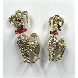 May include: A pair of gold-toned poodle brooches. Each brooch features a poodle with a red bow tie. The poodles have textured fur and black eyes. The brooches are set against a white background.