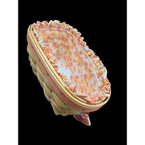 May include: A woven basket with a hinged lid. The basket is made of light-coloured wood with a reddish-orange trim. The interior features a fabric lining with a pattern of orange, yellow, and white candy corn. A decorative bow is attached to the front.