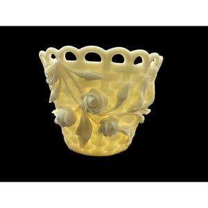 Puede incluir: Un recipiente decorativo en forma de cesta de color crema con una textura tejida. El recipiente está adornado con flores y hojas de rosas blancas. El borde de la cesta tiene un borde festoneado. El fondo es negro.