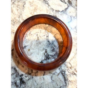 May include: A circular bangle bracelet featuring a polished, translucent amber-colored resin material with a smooth, rounded surface and a warm, variegated brown tone throughout the entire structure of the accessory.