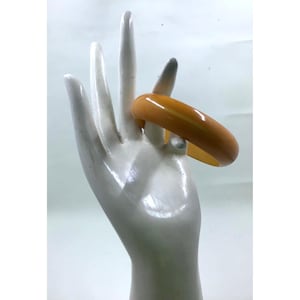 May include: A single, amber-colored bangle bracelet. The bracelet is a solid, smooth band with a glossy finish. The bracelet is displayed on a white hand-shaped display stand.